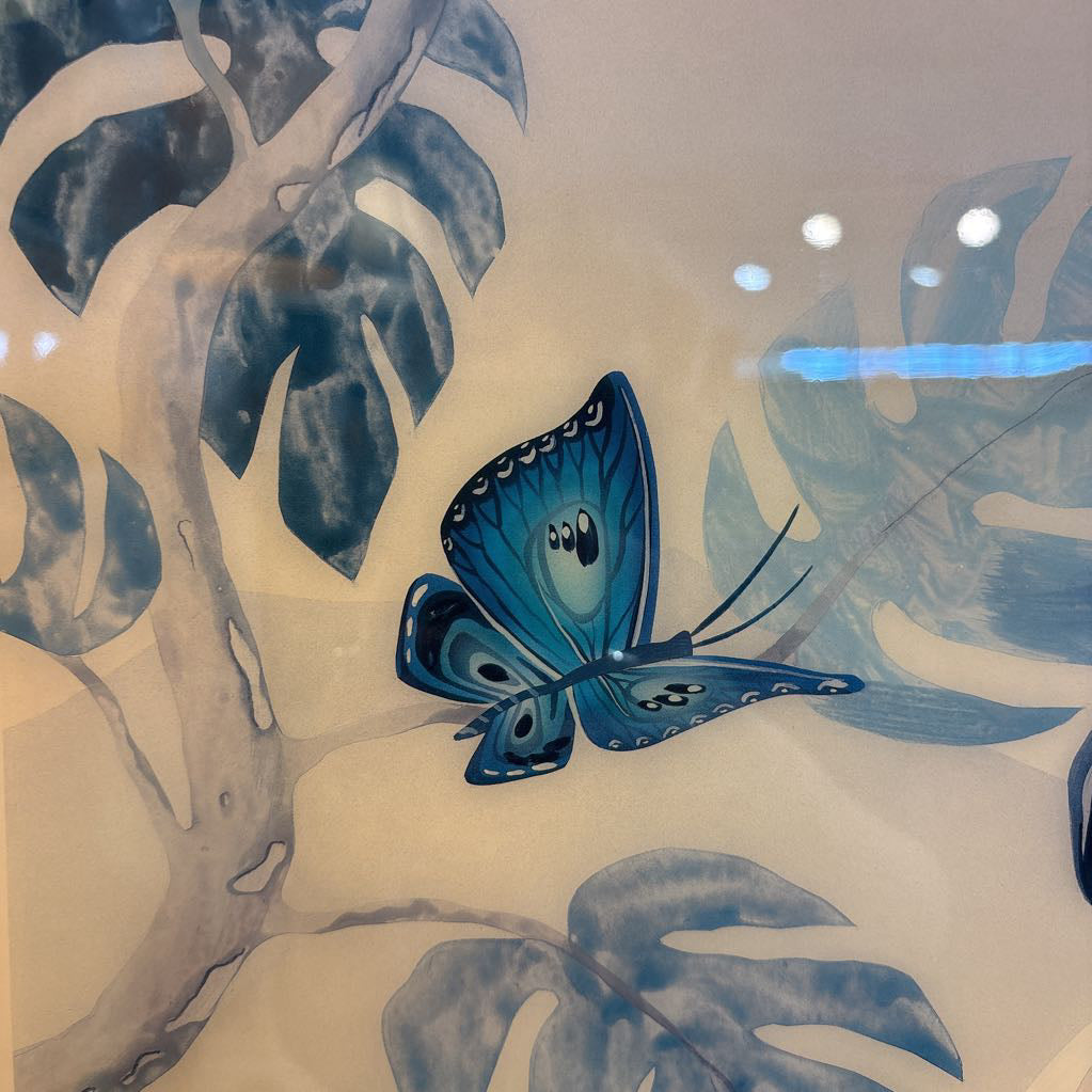 A close-up of a decorative surface featuring a blue butterfly perched among large blue and gray tropical leaves on a light beige background.