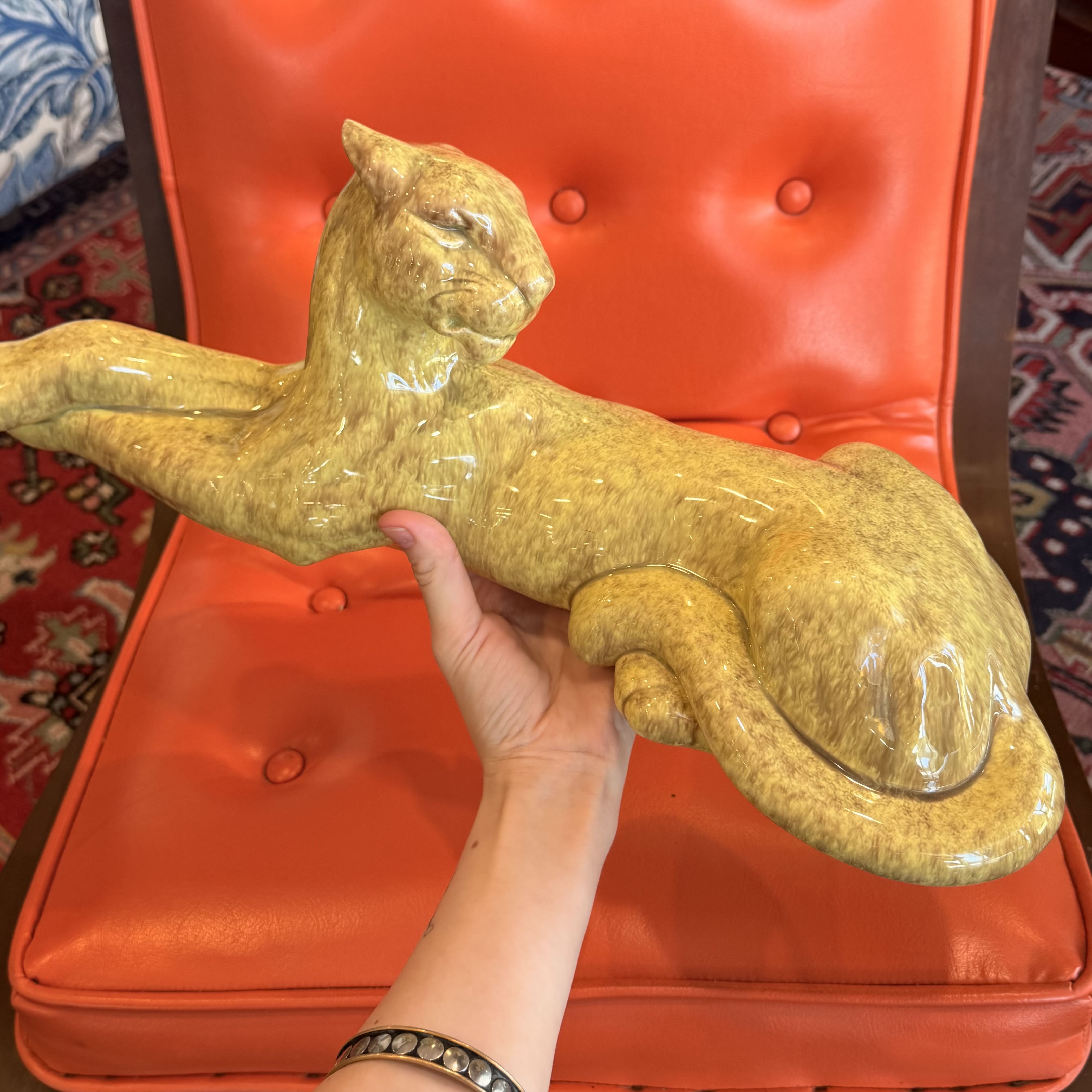 A glossy yellow-gold ceramic panther figurine being held by a person's hand in front of a bright orange tufted leather chair.