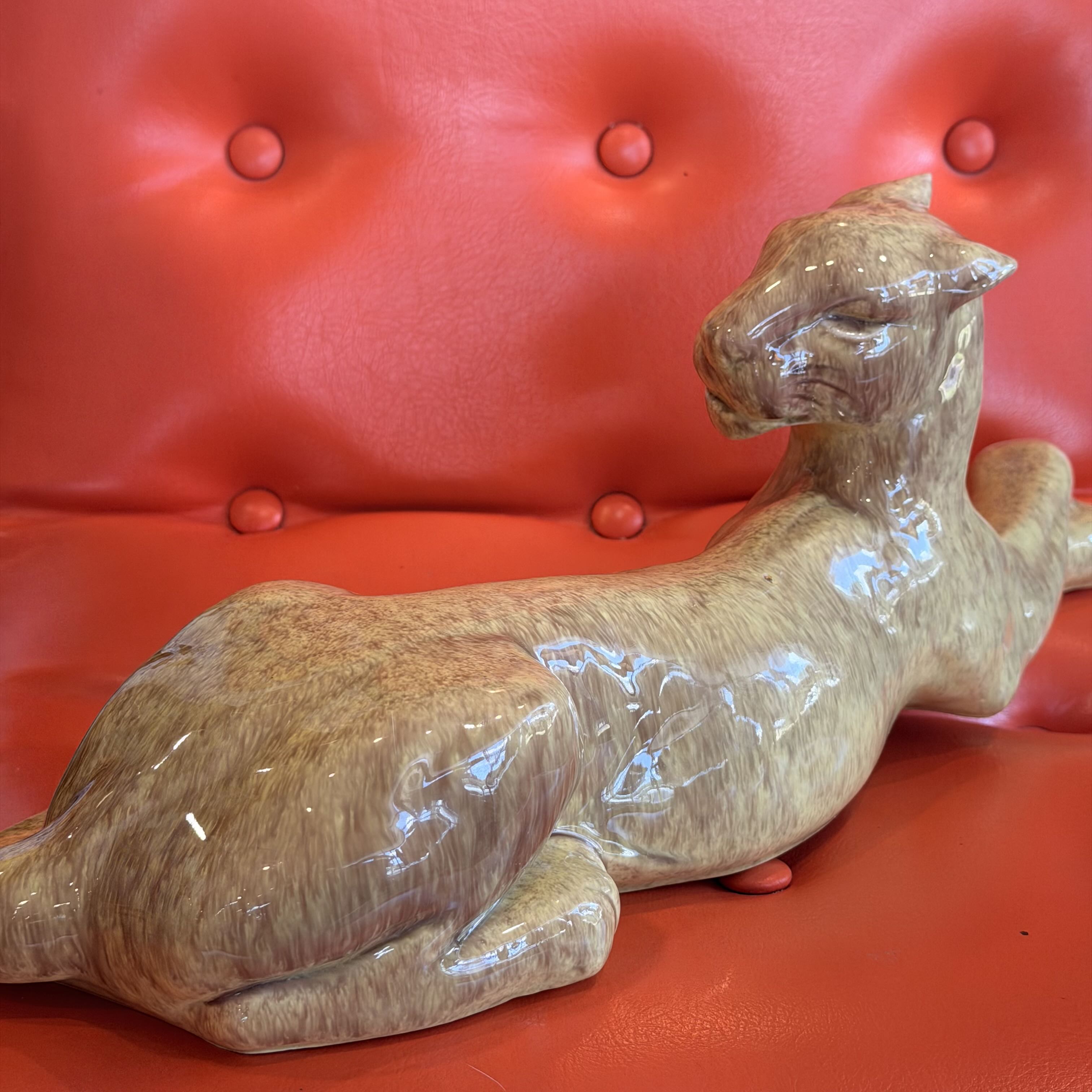 A glossy ceramic figurine of a reclining leopard or cheetah in warm tan and brown marbled tones, displayed on a bright red tufted leather sofa.