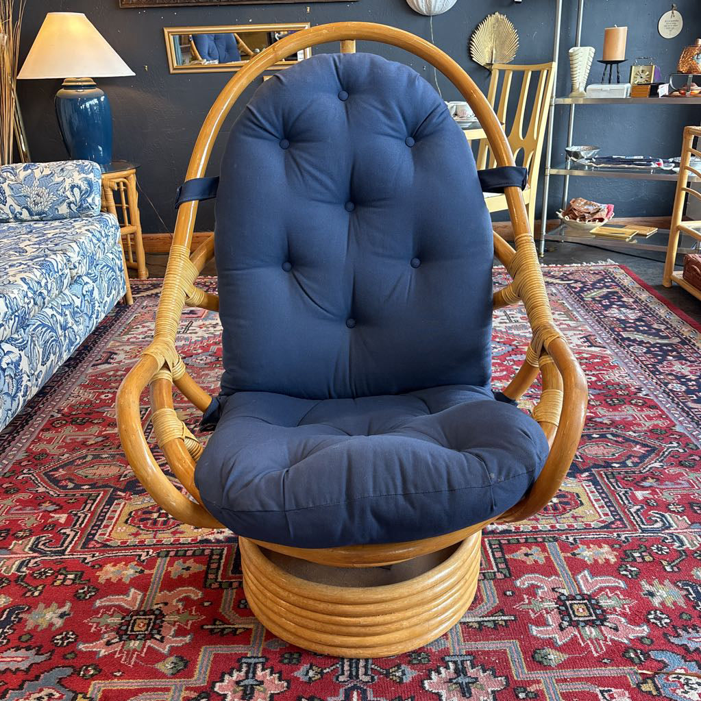 A rattan swivel armchair with a tall curved backrest and thick tufted navy blue seat and back cushions, shown in a furnished room on a patterned red rug.