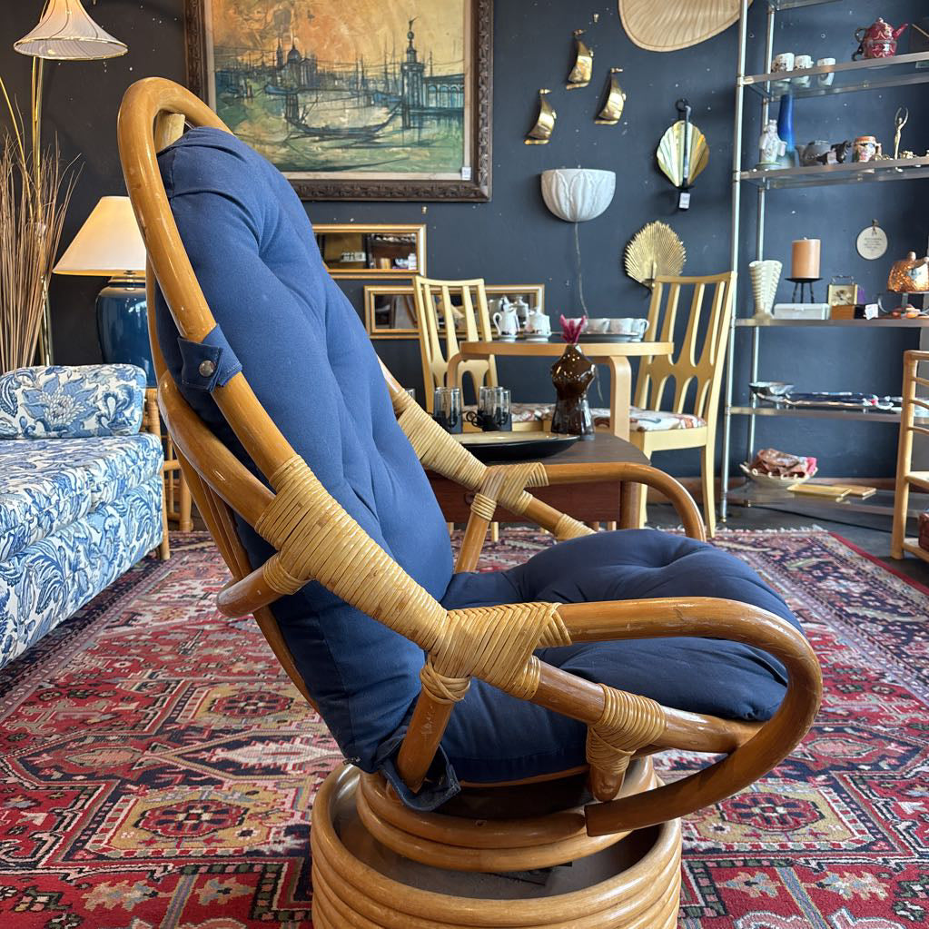 A mid-century modern rattan swivel armchair with thick navy blue cushions, set in a rustic, cluttered home decor showroom.