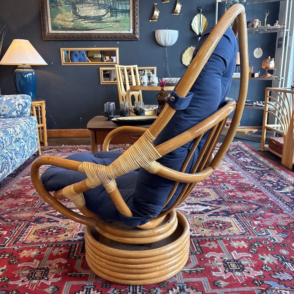A tall, curved rattan rocking chair with a high back, wrapped in natural woven material, and upholstered with dark blue cushions, shown in a vintage-style room with a patterned red rug and dark blue wall.