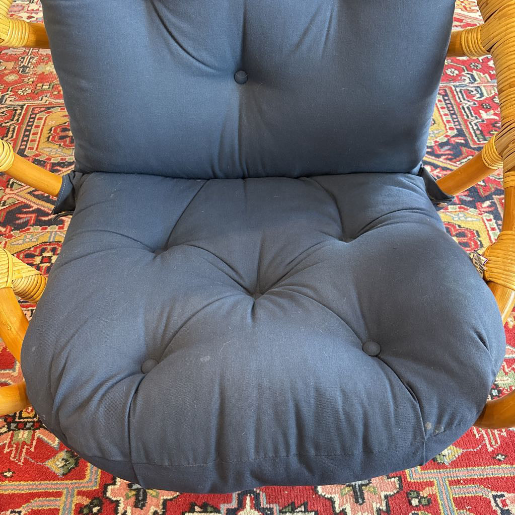 A close-up of a tufted blue-gray upholstered chair cushion, showing the seat and backrest, with a woven rattan frame and a red patterned rug underneath.