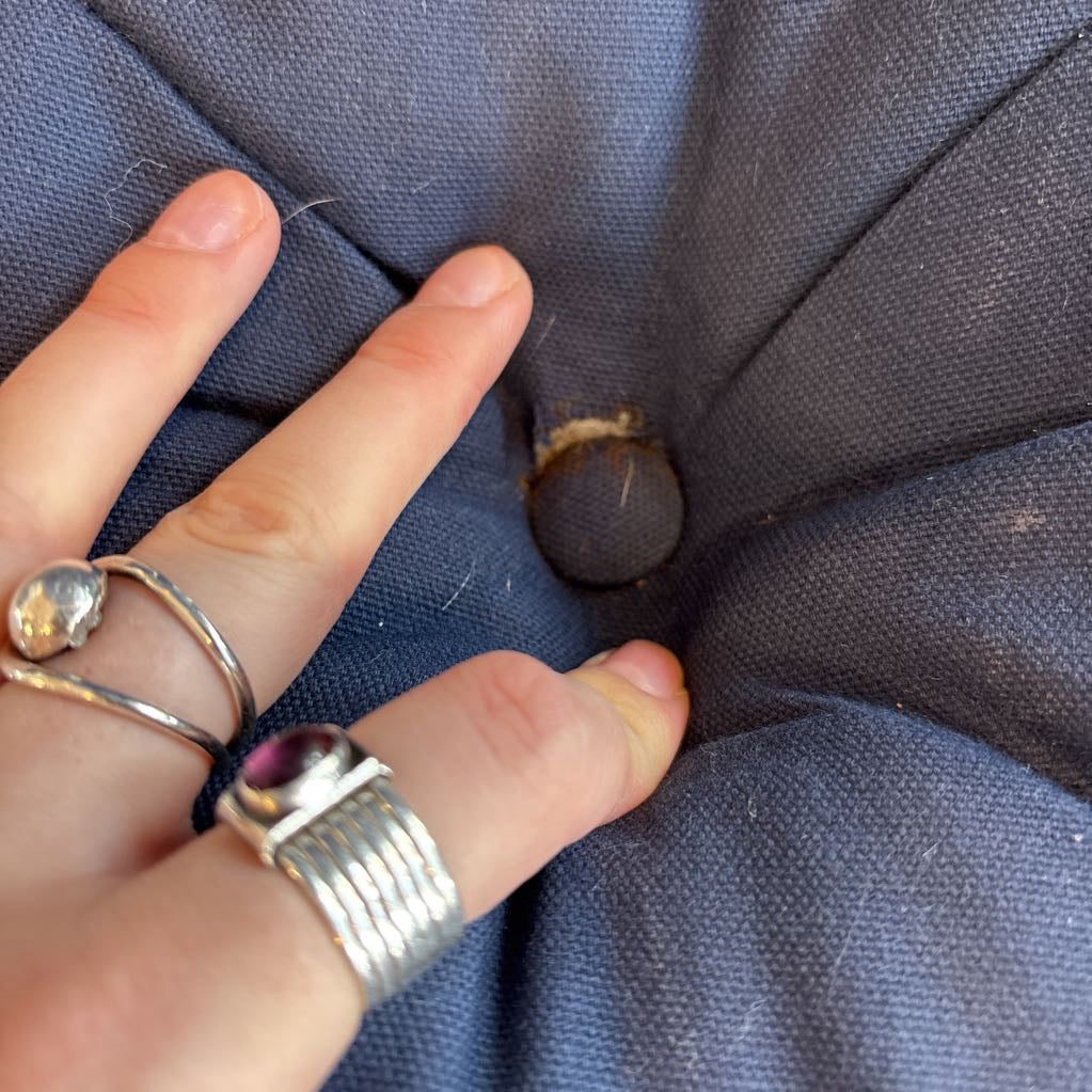 A close-up of a hand pressing into a tufted button on a dark blue fabric piece, wearing silver rings.