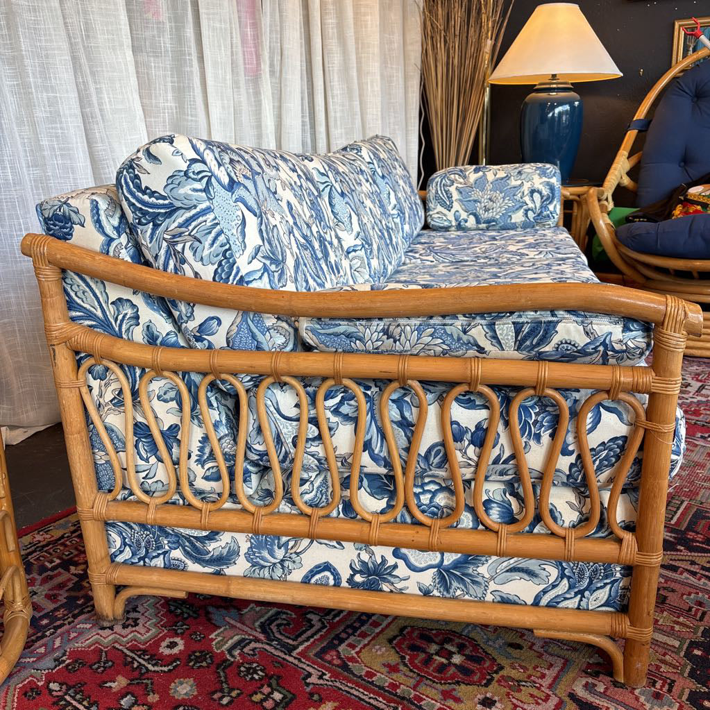 A rattan loveseat sofa with blue and white toile floral pattern cushions, shown in a home setting on a patterned rug.