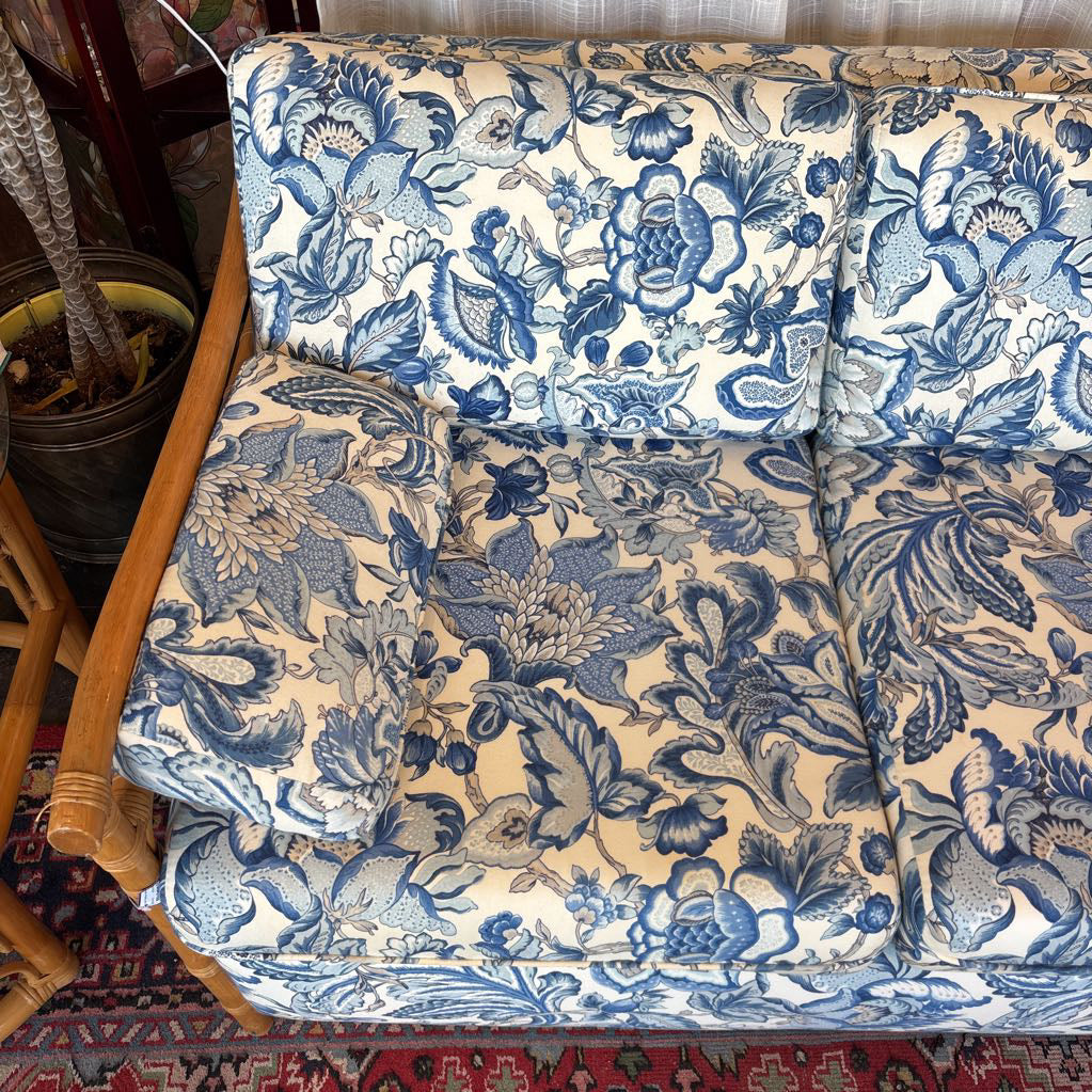 A close-up of a blue and cream floral toile patterned fabric on a sofa or chair, with visible wooden arms and part of a patterned rug underneath.