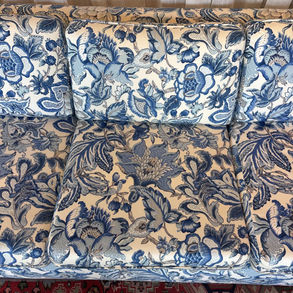 Blue and beige floral upholstery fabric shown on a sofa cushion set, featuring a detailed botanical pattern with blue flowers and leaves on a warm cream background.