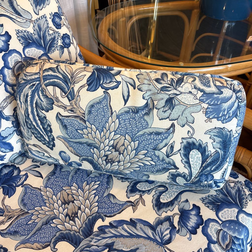 Blue and white floral patterned outdoor chair cushion on a rattan chair, with a matching cushion resting on the seat. A glass-top table is visible in the background.