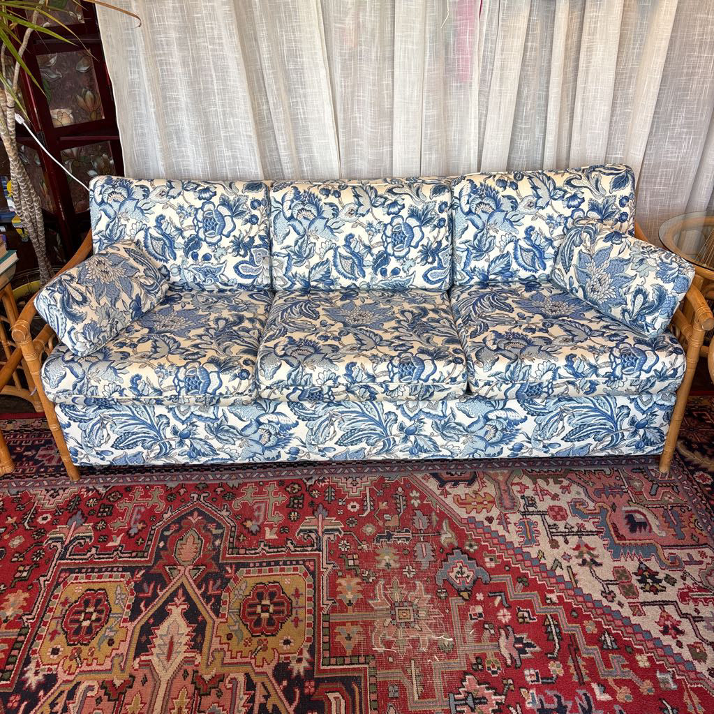 A three-seat sofa upholstered in a blue and cream floral toile-style pattern, set on a red patterned oriental rug in front of sheer curtains.