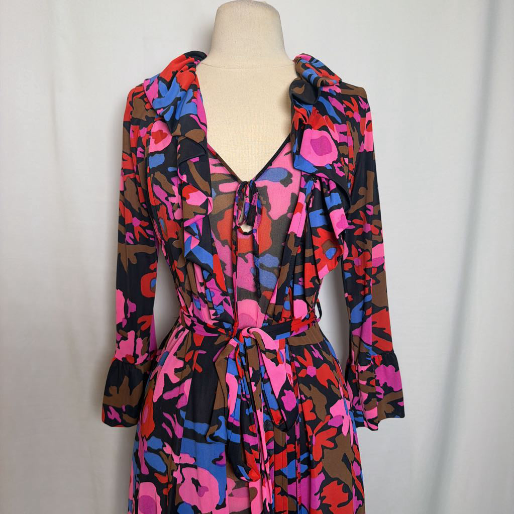 A long-sleeve wrap dress with a vibrant abstract floral pattern in pink, blue, red, and brown on a black background, displayed on a cream dress form against a light gray fabric backdrop.