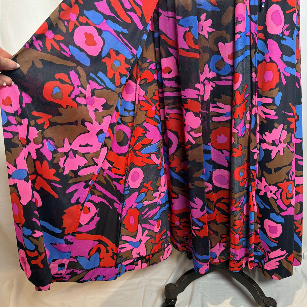 A pleated fabric garment or scarf with a bold abstract floral pattern in pink, red, blue, and brown on a black background, held open by a hand against a white fabric backdrop.