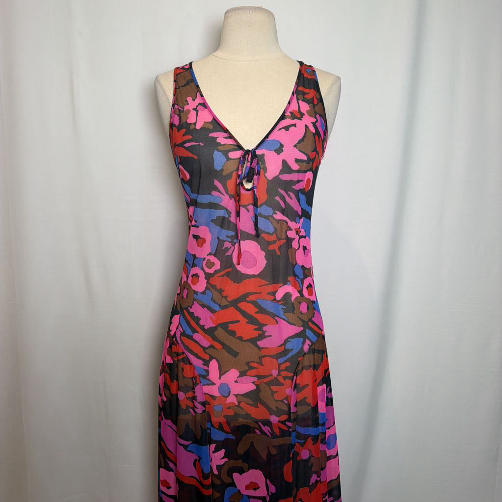 A sleeveless V-neck dress with a tie-front detail, featuring a vibrant abstract floral pattern in pink, red, blue, and brown over a sheer black mesh fabric, displayed on a mannequin torso against a white curtain backdrop.