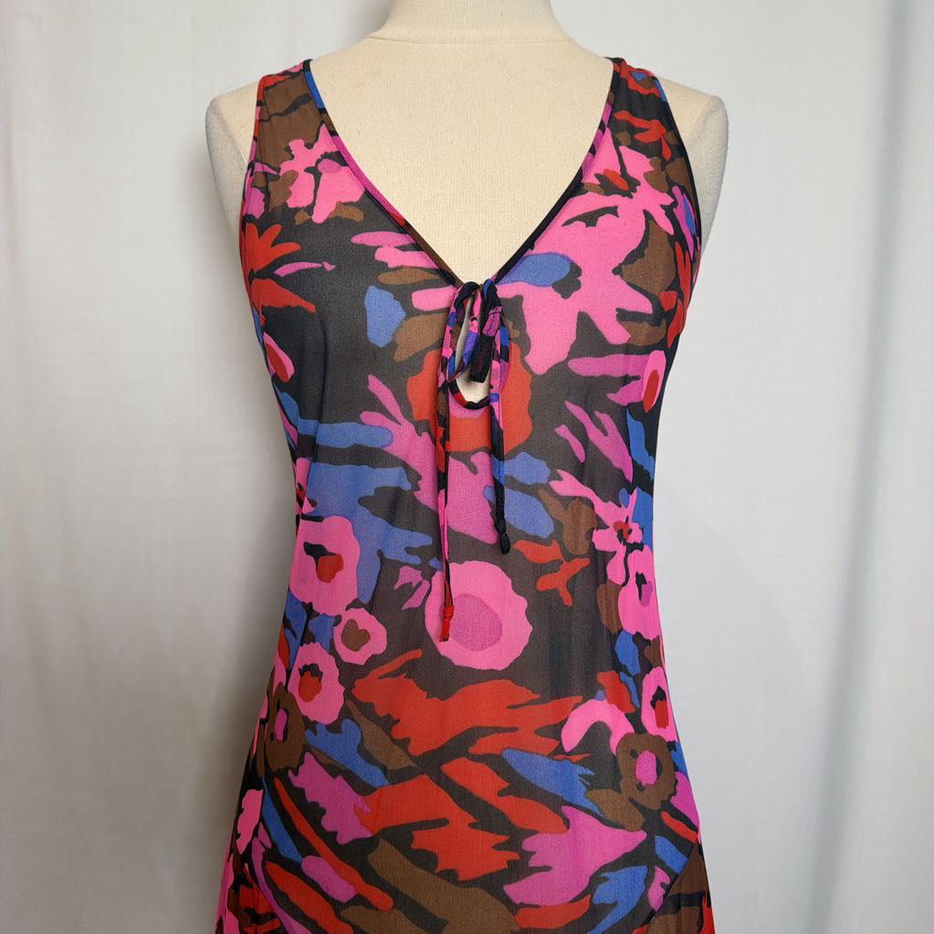 A sleeveless V-neck tie-front dress displayed on a cream dress form against a light gray fabric backdrop. The dress features a bold abstract floral pattern in pink, red, blue, and brown over a sheer black mesh fabric.