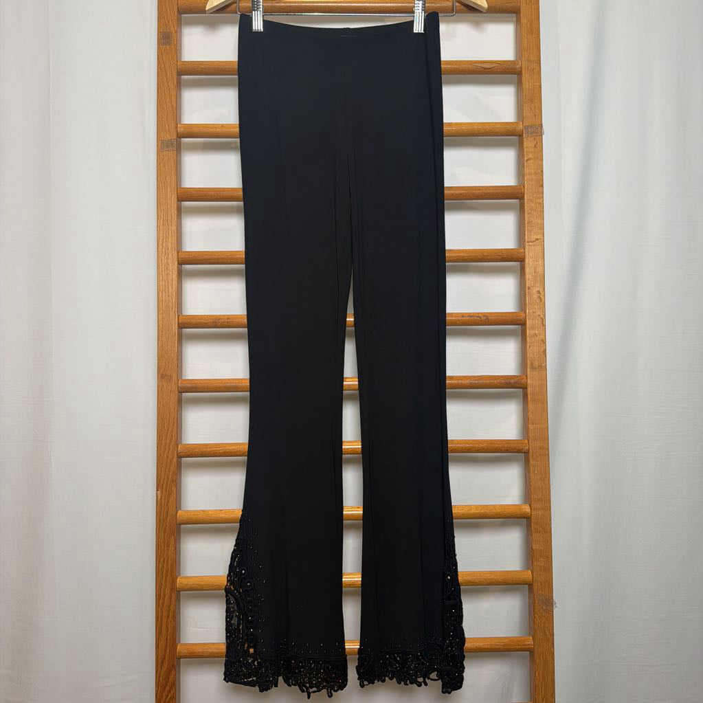 Black wide-leg pants with a lace hem, hanging on a wooden ladder rack against a white curtain backdrop.