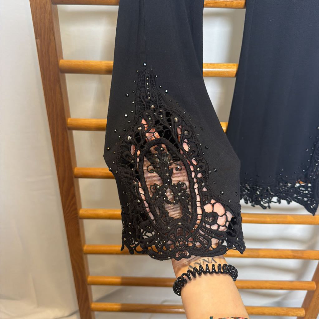 Black wide-leg pants with intricate lace and bead trim at the hems, shown hanging on a wooden rack with a person's hand visible.