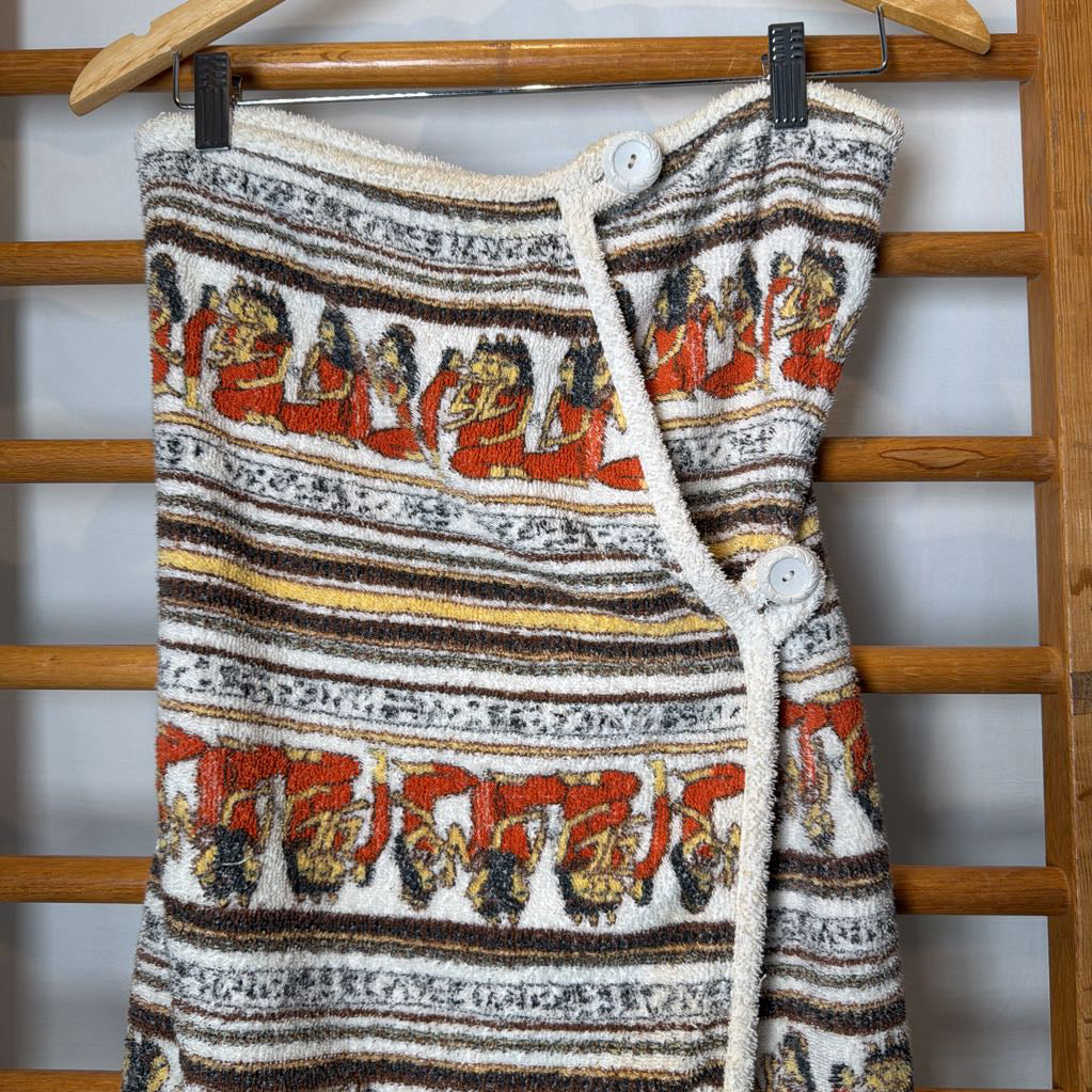 A textured knit wrap dress with a cream base and horizontal stripes in brown, black, and gold, featuring red and orange figures woven into the pattern. The dress has a cream rope tie closure and is shown hanging on a wooden hanger against a wooden slatted background.