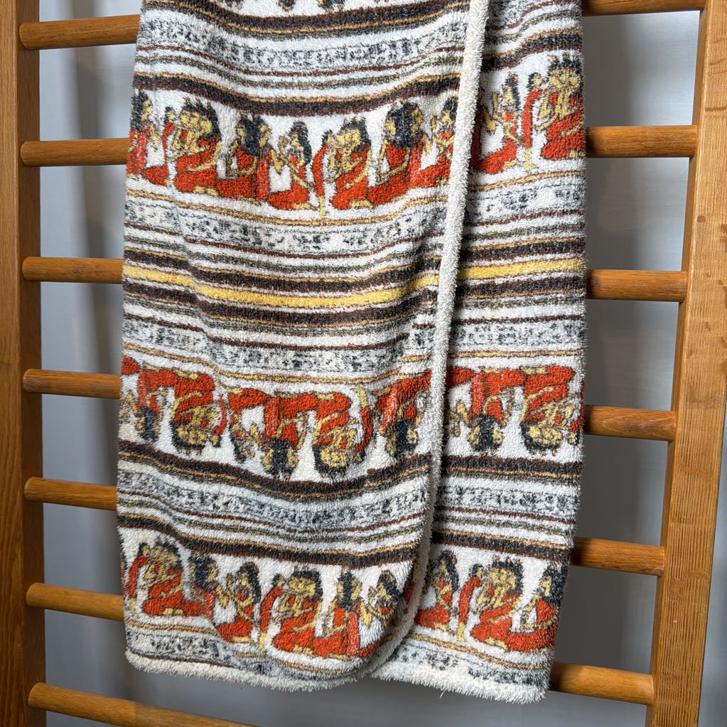 A textured knit blanket with a repeating horizontal pattern of figures in red, brown, and cream, draped over a wooden towel rack.