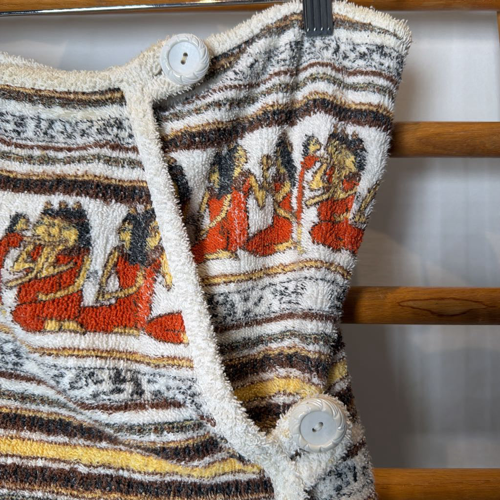 A close-up of a textured knit garment featuring horizontal stripes in cream, brown, black, and yellow, with a central panel of orange and red figures. The garment has a cream tassel trim and two large white buttons, hanging against a wooden slatted background.
