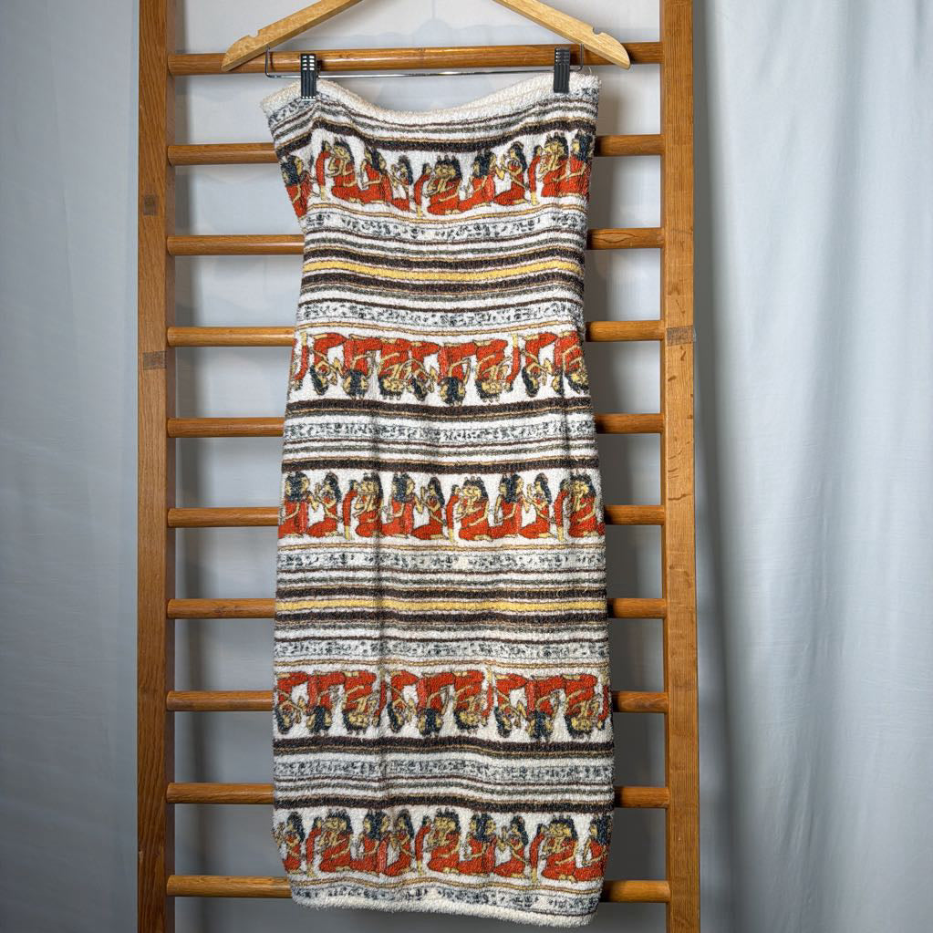 A sleeveless knit dress with horizontal bands of black, white, orange, and brown, featuring a repeating pattern of stylized faces, hanging on a wooden hanger from a wooden ladder against a light gray backdrop.
