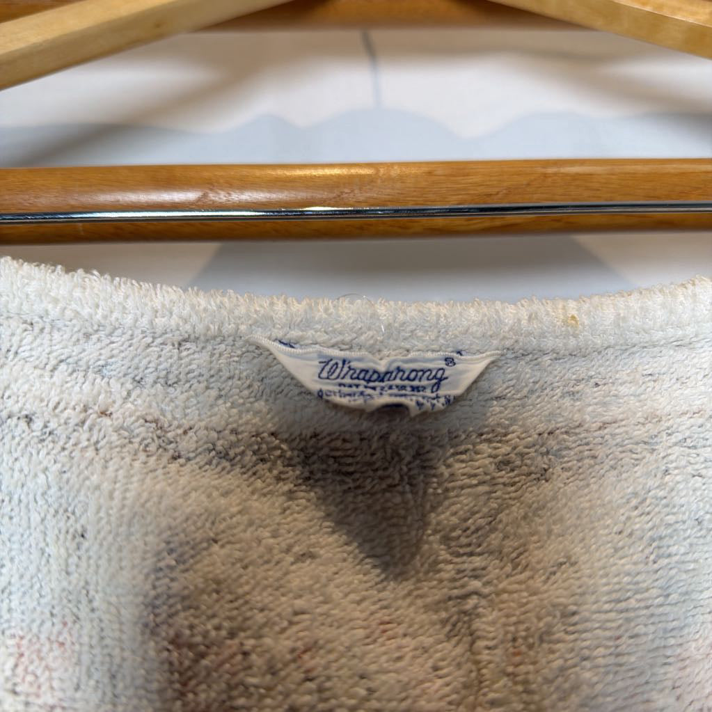 Close-up of a white terry cloth towel hanging on a wooden hanger, showing the brand label 'Wrapphnary' sewn into the neck.
