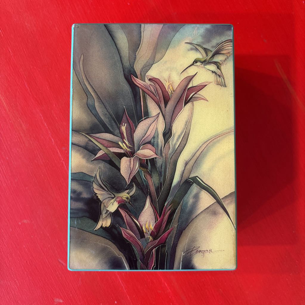 A rectangular decorative box or package featuring a botanical illustration with pink and cream flowers, green leaves, and a hummingbird, photographed against a bright red background.