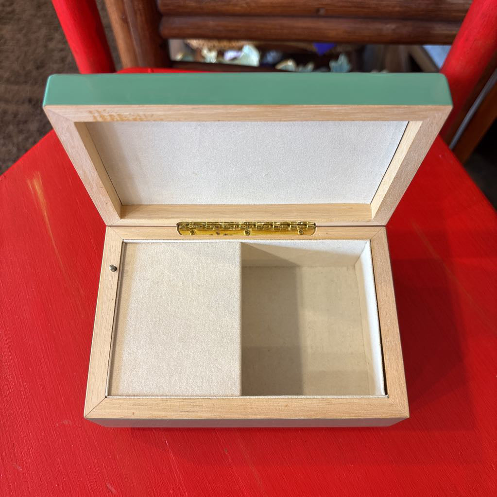 An open wooden jewelry box with a light natural wood frame, a mint green lid, and a beige velvet interior, shown on a bright red surface.