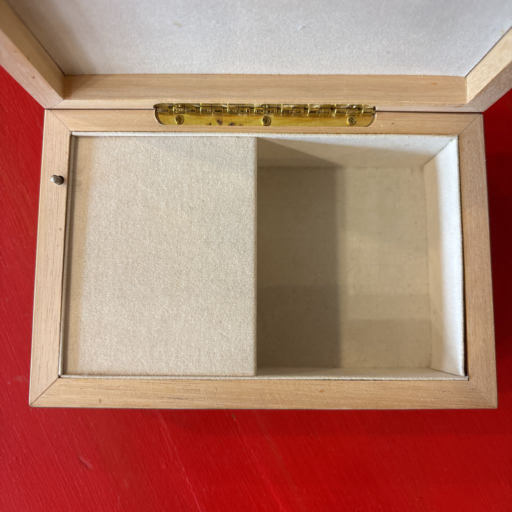 An open wooden jewelry box with a light natural wood frame, a mint green lid, and a beige velvet interior, shown on a bright red surface.
