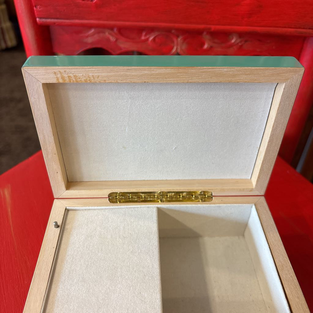 An open wooden jewelry box with a light natural wood frame, a mint green lid, and a beige velvet interior, shown on a bright red surface.