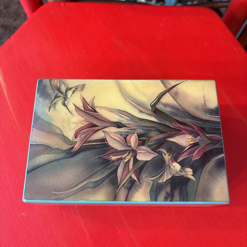 A rectangular decorative box or package featuring a botanical illustration with pink and cream flowers, green leaves, and a hummingbird, photographed against a bright red background.