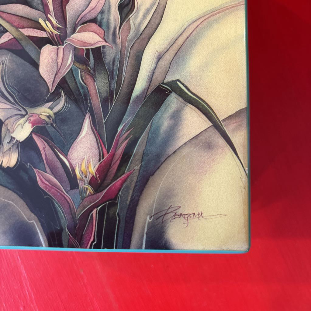 A rectangular decorative box or package featuring a botanical illustration with pink and cream flowers, green leaves, and a hummingbird, photographed against a bright red background.