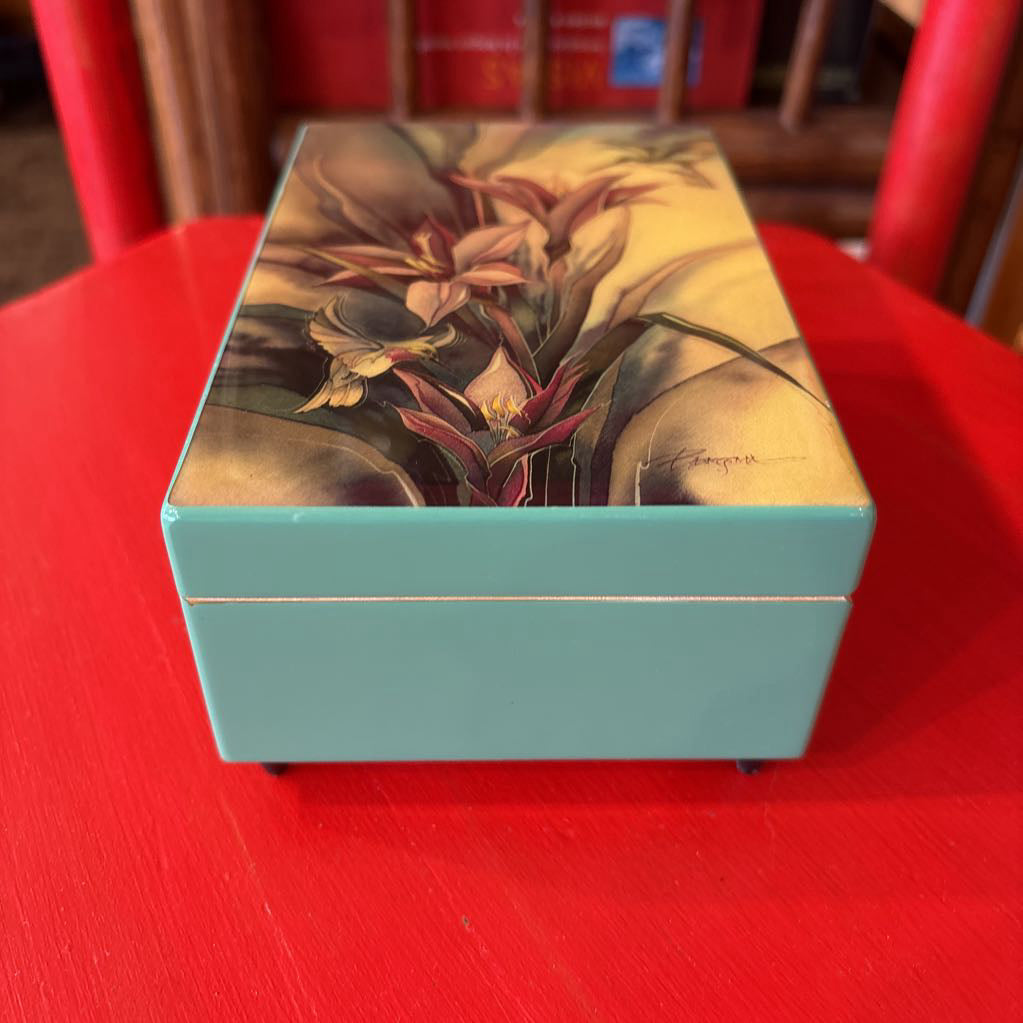 A rectangular lacquered box with a light blue base and a floral lid design, placed on a red table in front of a red chair.