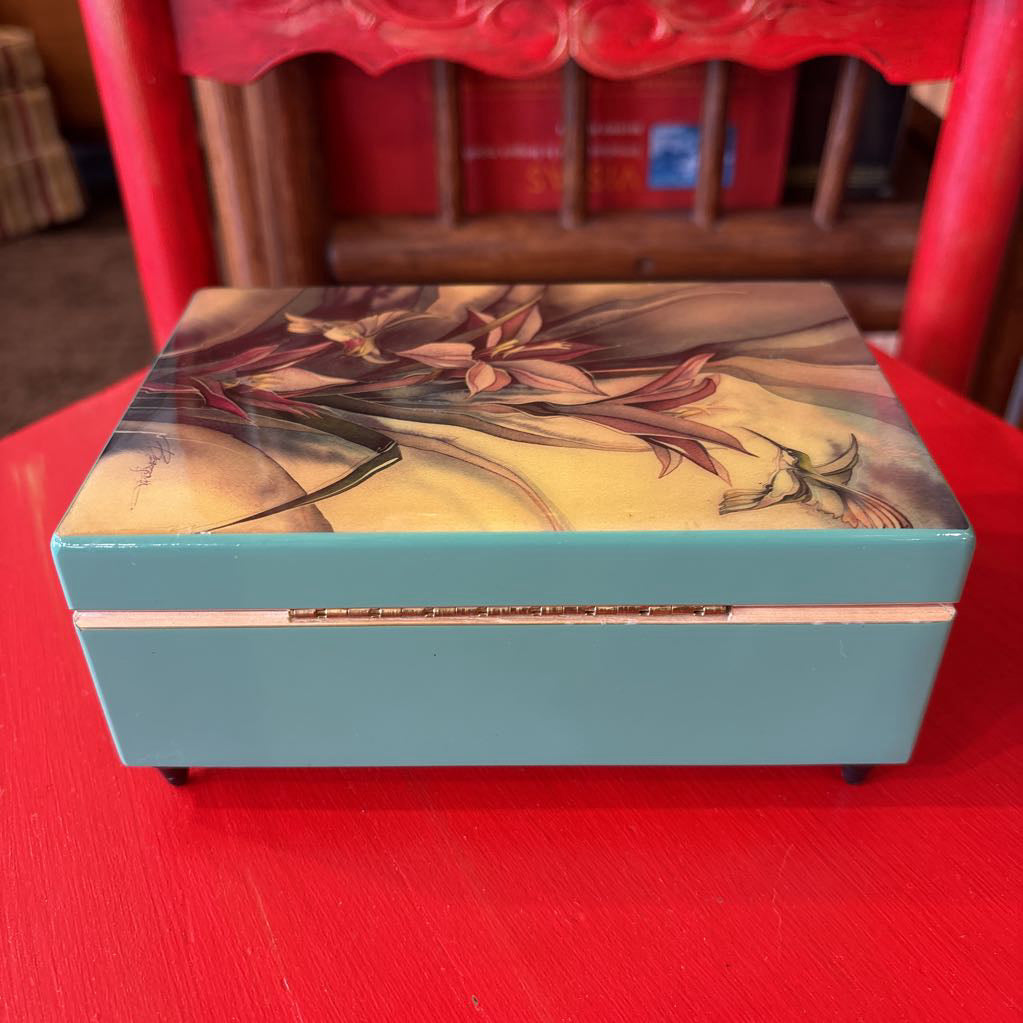 A rectangular lacquered box with a light blue base and a floral lid design, placed on a red table in front of a red chair.