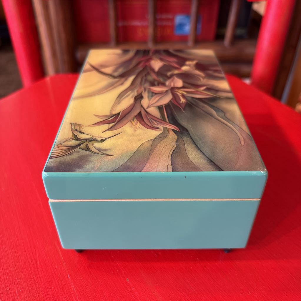 A rectangular turquoise blue box with a floral lid design, placed on a bright red table with a blurred indoor background.