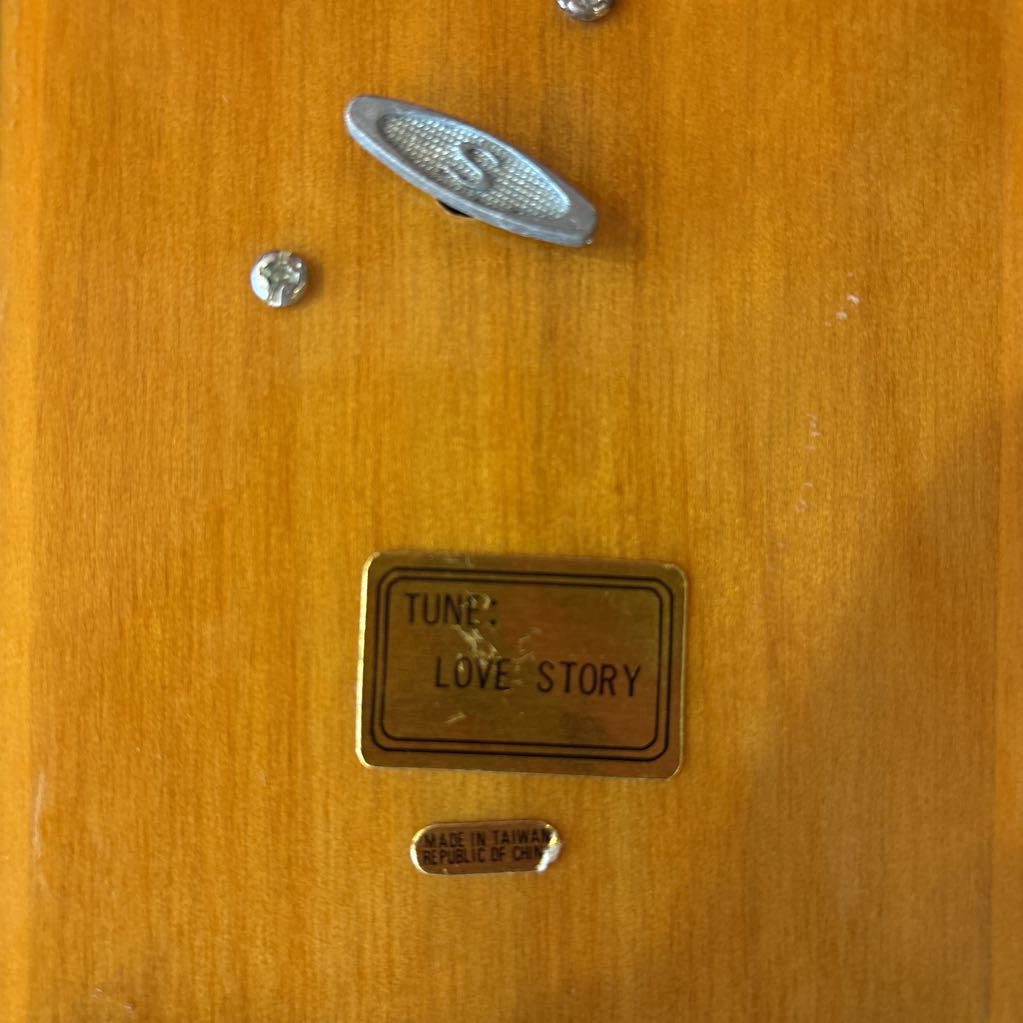 The bottom of wooden music box with a crank and a sticker that says 'TUNE: LOVE STORY' and 'Made in Taiwan Republic of China.'