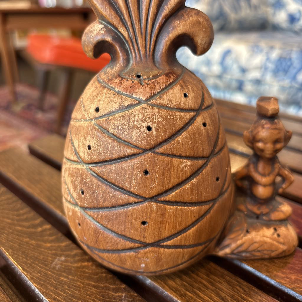 A wooden pineapple and small sitting figure toothpick holder on a wooden bench in a room setting.