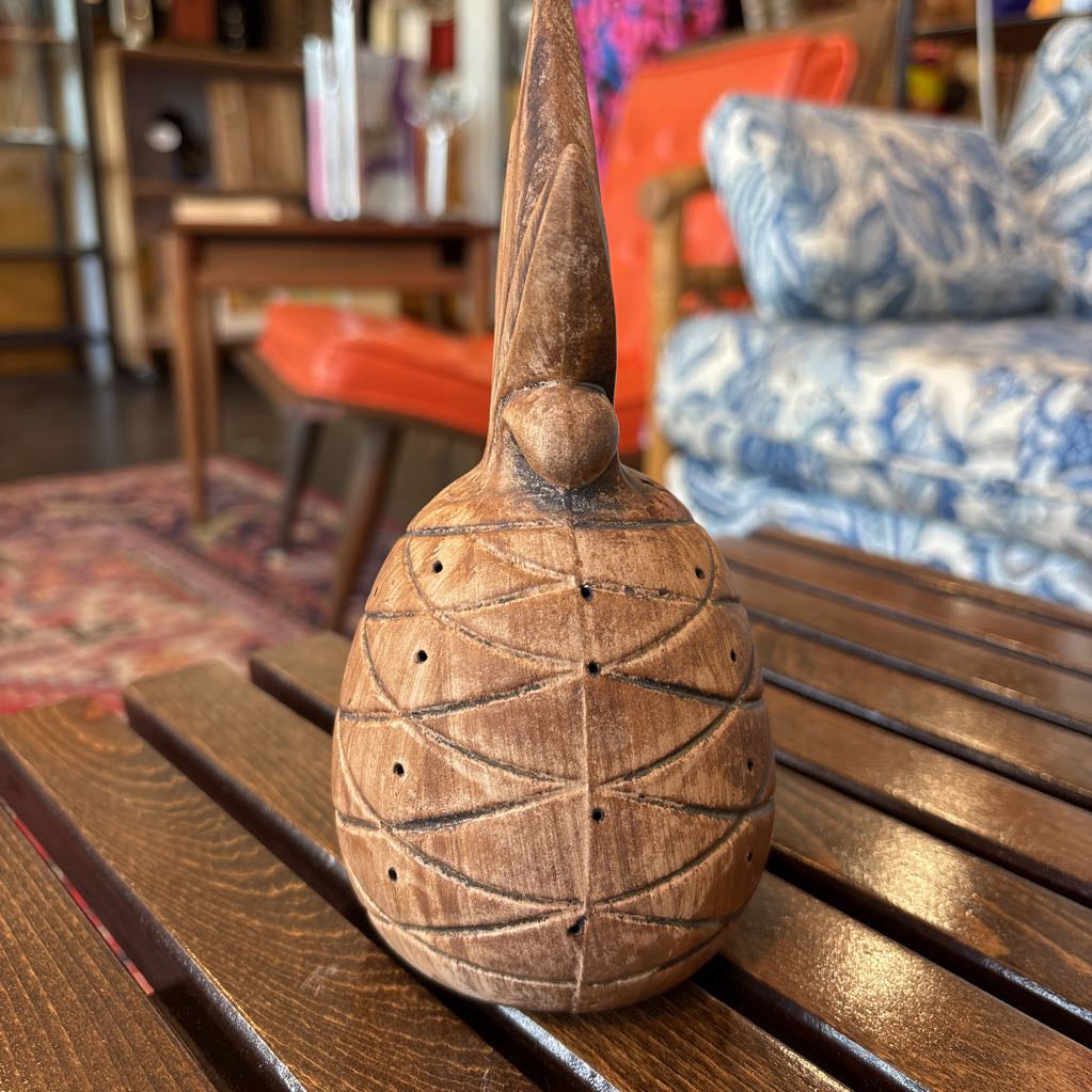 A wooden pineapple and small sitting figure toothpick holder on a wooden bench in a room setting.