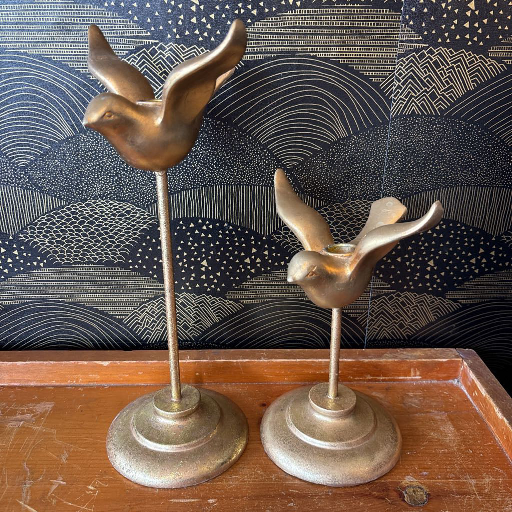 Two vintage brass bird candlesticks shaped like flying doves, displayed on a wooden surface against a dark patterned wallpaper background.