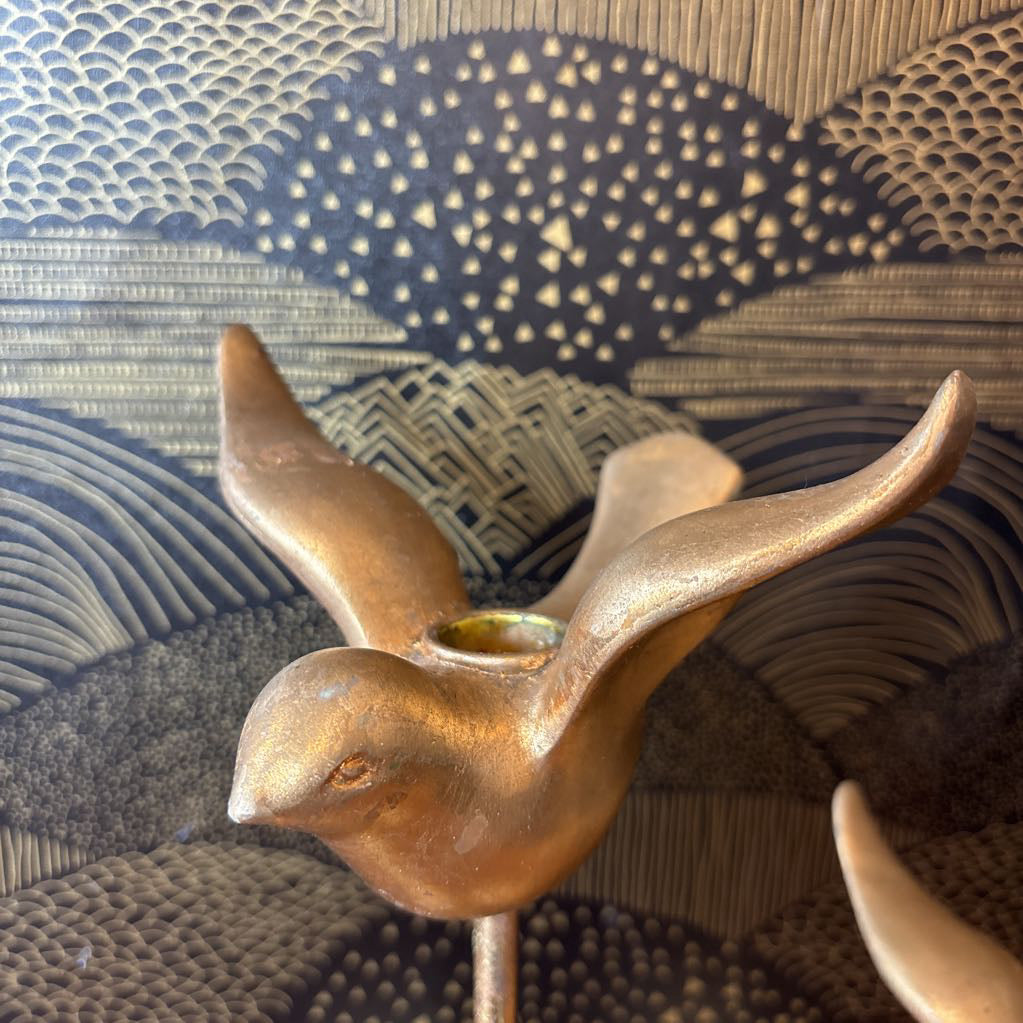 A close-up of a metallic bird-shaped object with wings spread, possibly a decorative sculpture or candle holder, shown against a patterned woven background.