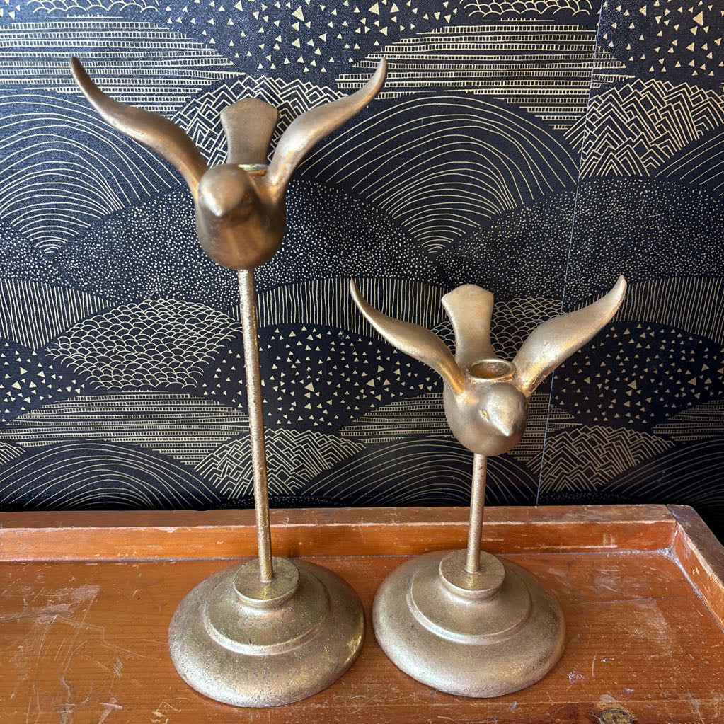 Two vintage brass bird-shaped candle holders displayed on a wooden surface against a dark patterned backdrop.