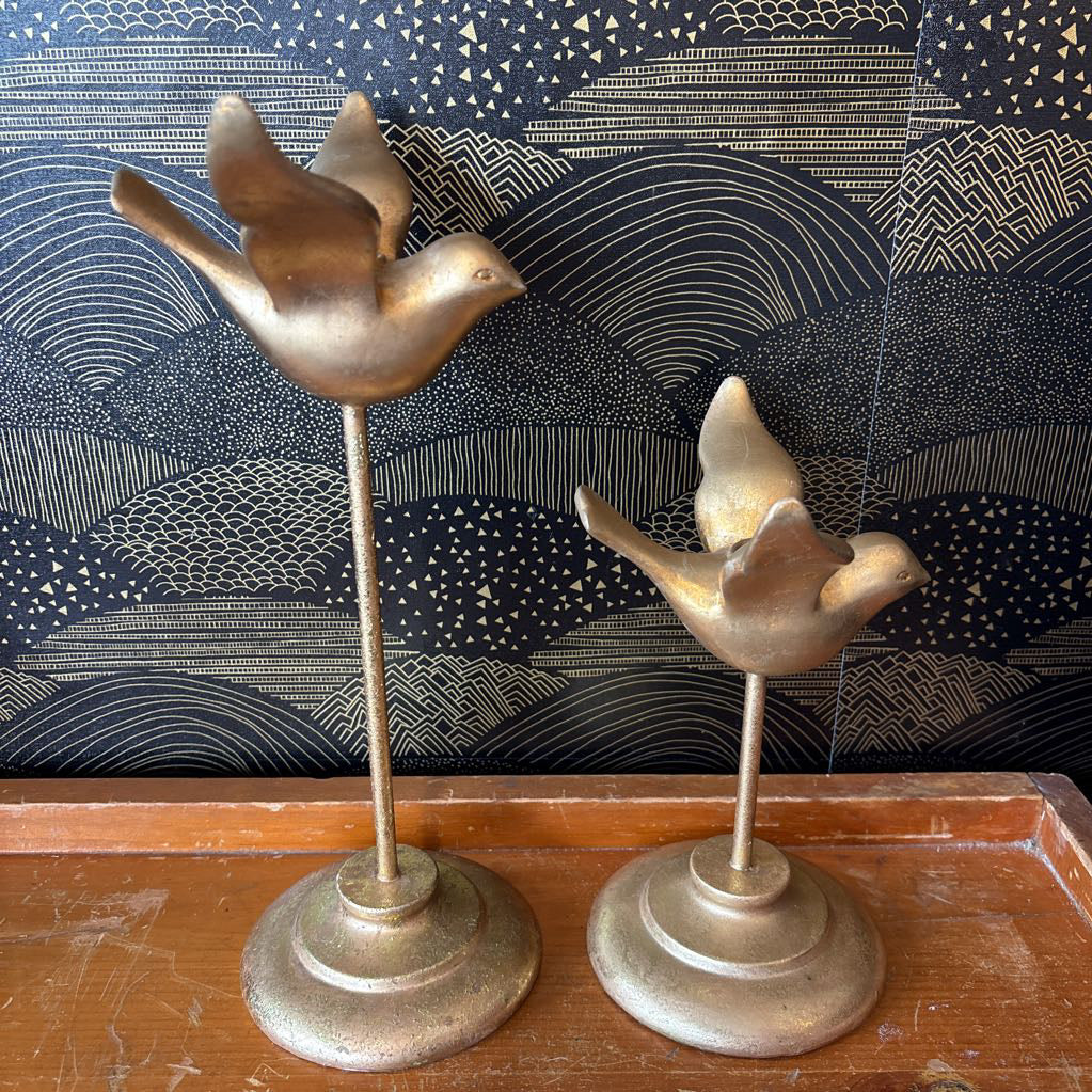 Two vintage-style brass bird candlesticks on a wooden surface, set against a dark patterned wallpaper background.