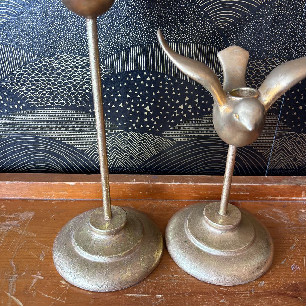 Two vintage brass bird-shaped candlesticks on a wooden table against a patterned wallpaper background.