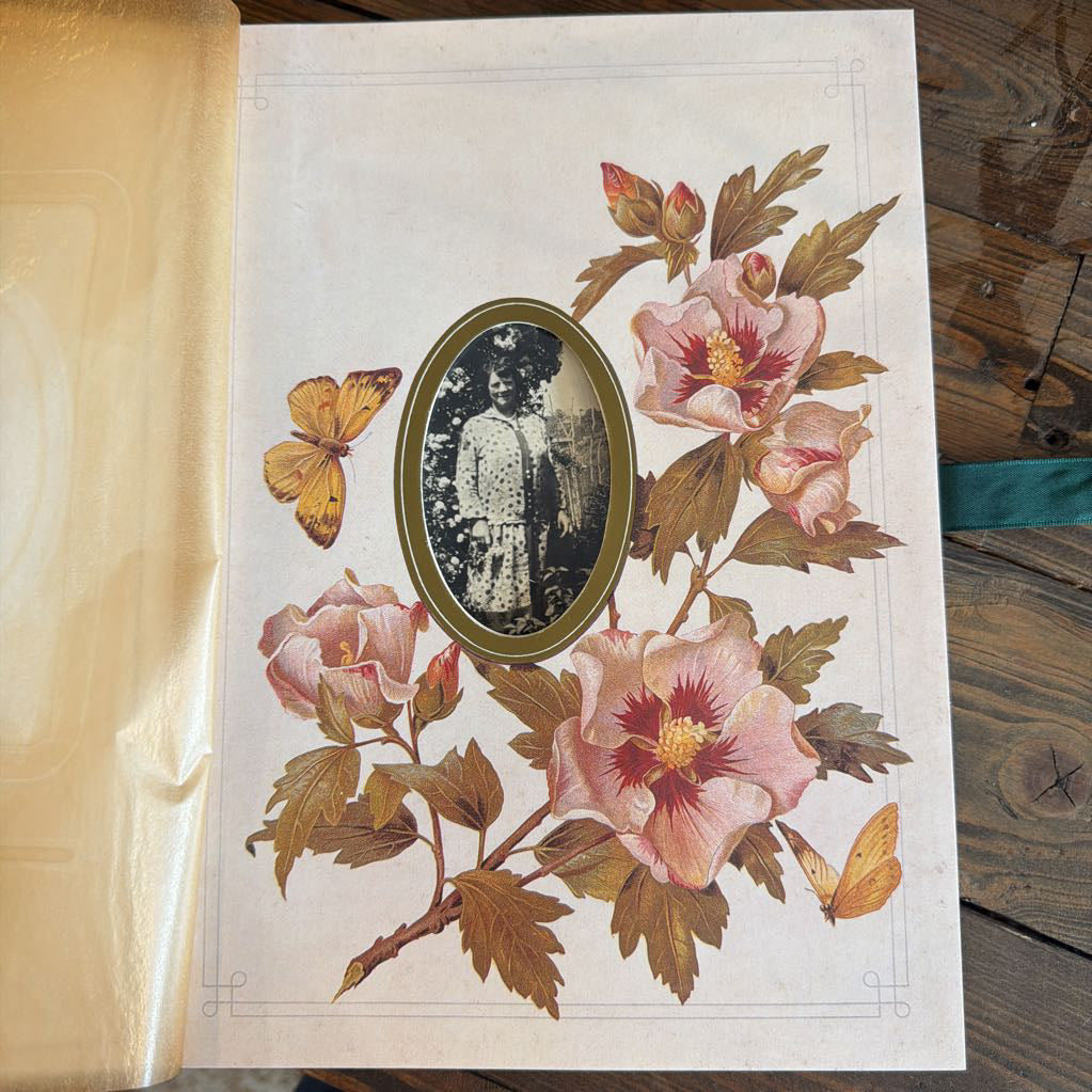An antique-style printed card or page with a vintage floral illustration, including pink flowers, brown leaves, yellow butterflies, and an oval inset portrait, photographed on a wooden surface next to a gold-toned envelope.
