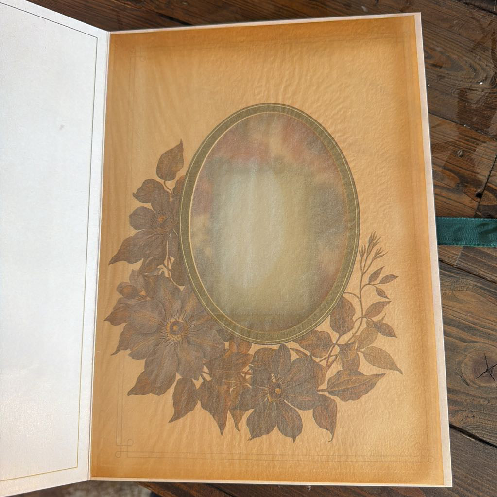 An open vintage-style book or album with a tan interior page featuring an oval portrait frame surrounded by floral illustrations, photographed from above on a wooden surface.