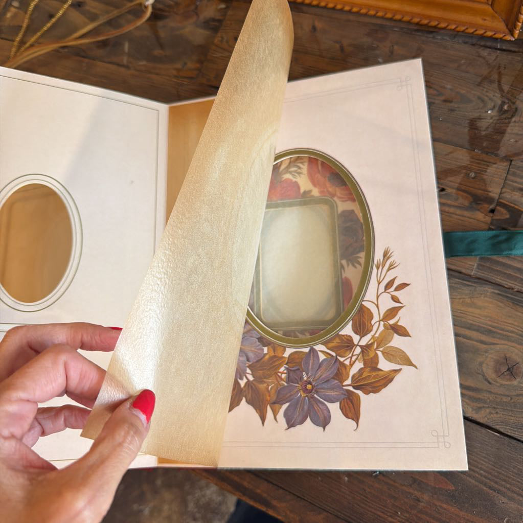 A hand with red nail polish holds a beige textured paper sheet over an open vintage-style book or album with floral illustrations and oval photo slots on the pages, set on a wooden table.