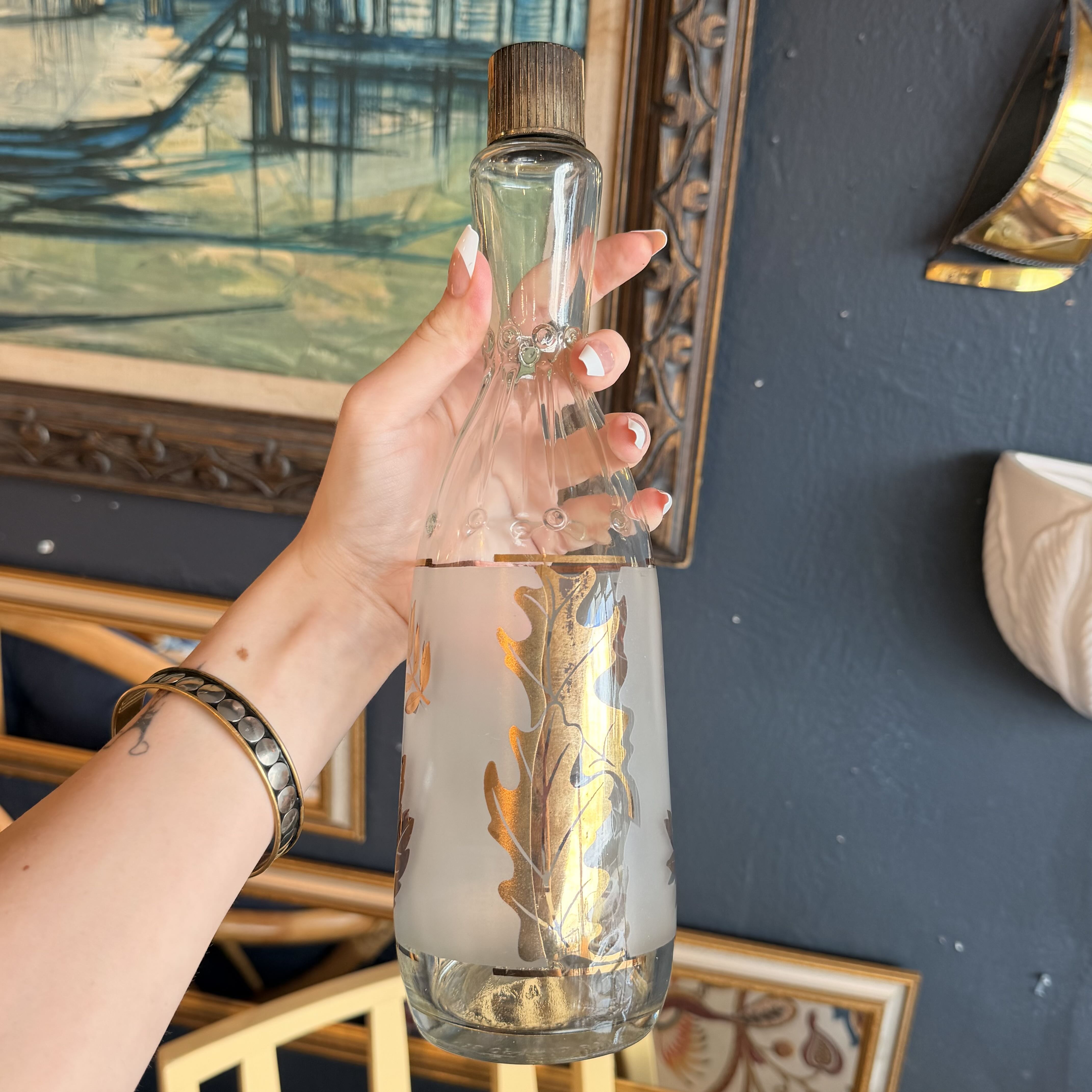 A hand holds a frosted glass bottle with a gold leaf pattern and a dark brown wooden cap, set against a dark blue wall with framed artwork.