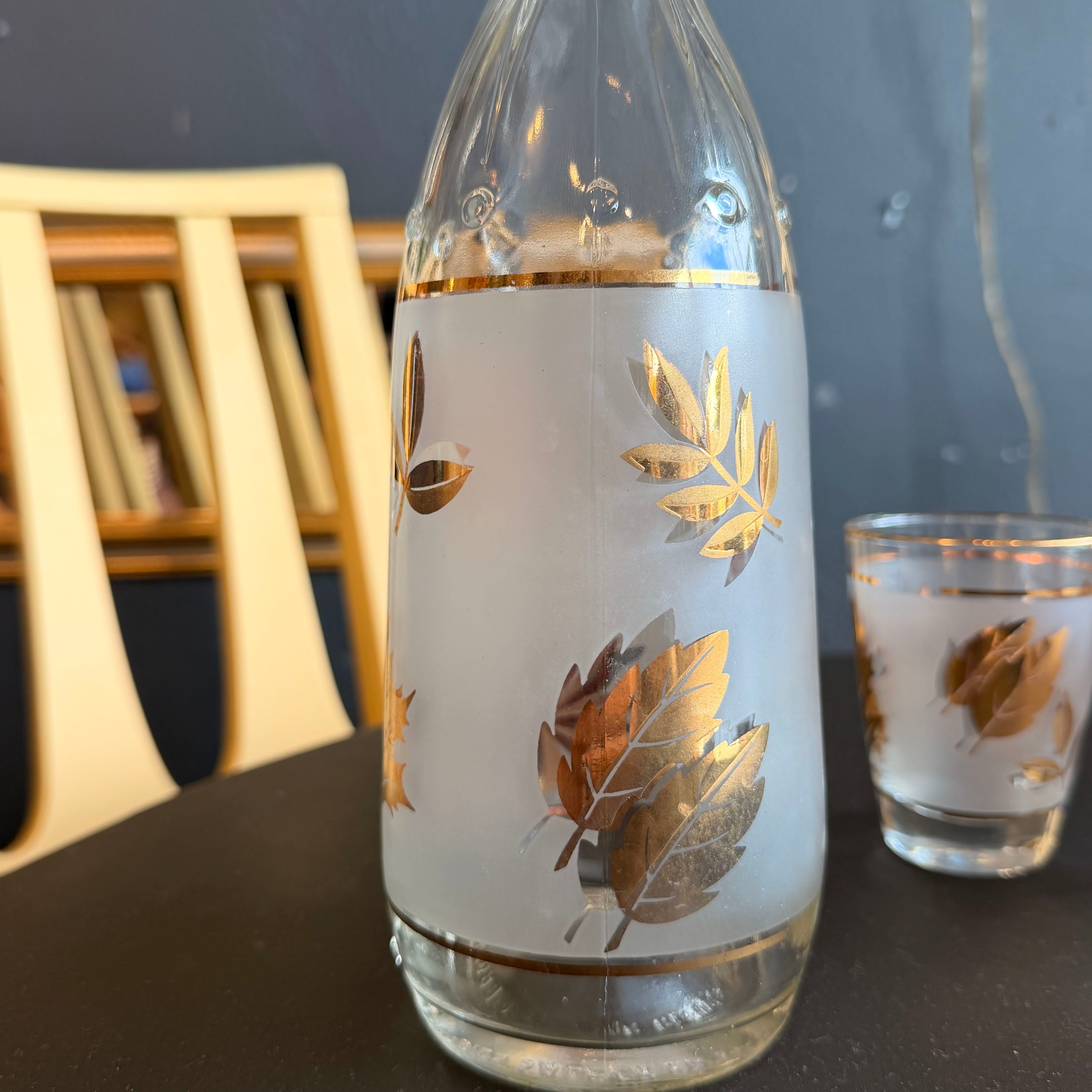 A frosted glass sake bottle with metallic gold leaf designs, shown alongside a matching small glass on a dark tabletop in an indoor setting.