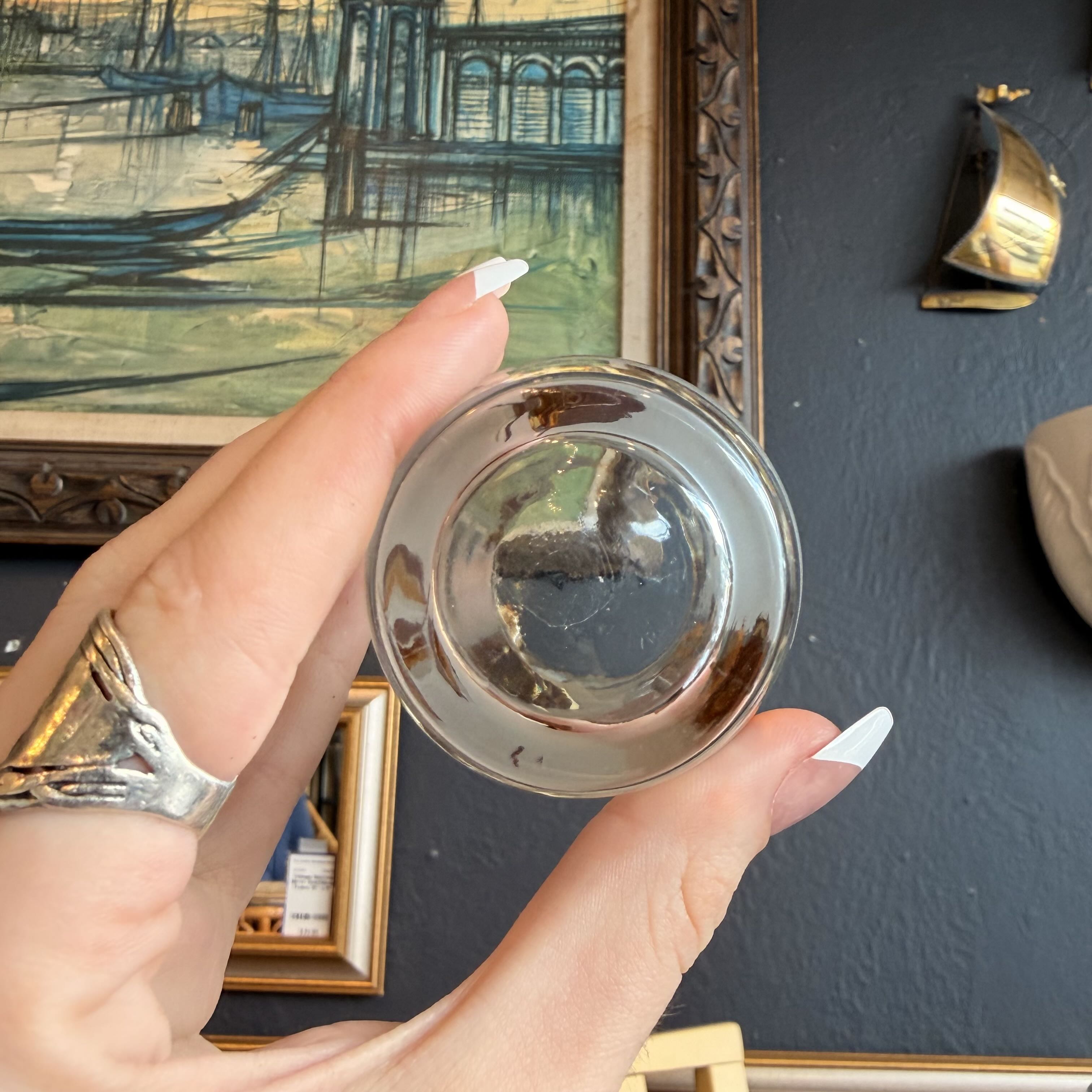 A hand with white manicured nails holds a small, clear glass bowl or dish, showing its reflective, dome-like interior. The background features a dark gray wall with framed artwork, including a vintage-style painting of a harbor and a gold-toned decorative piece.
