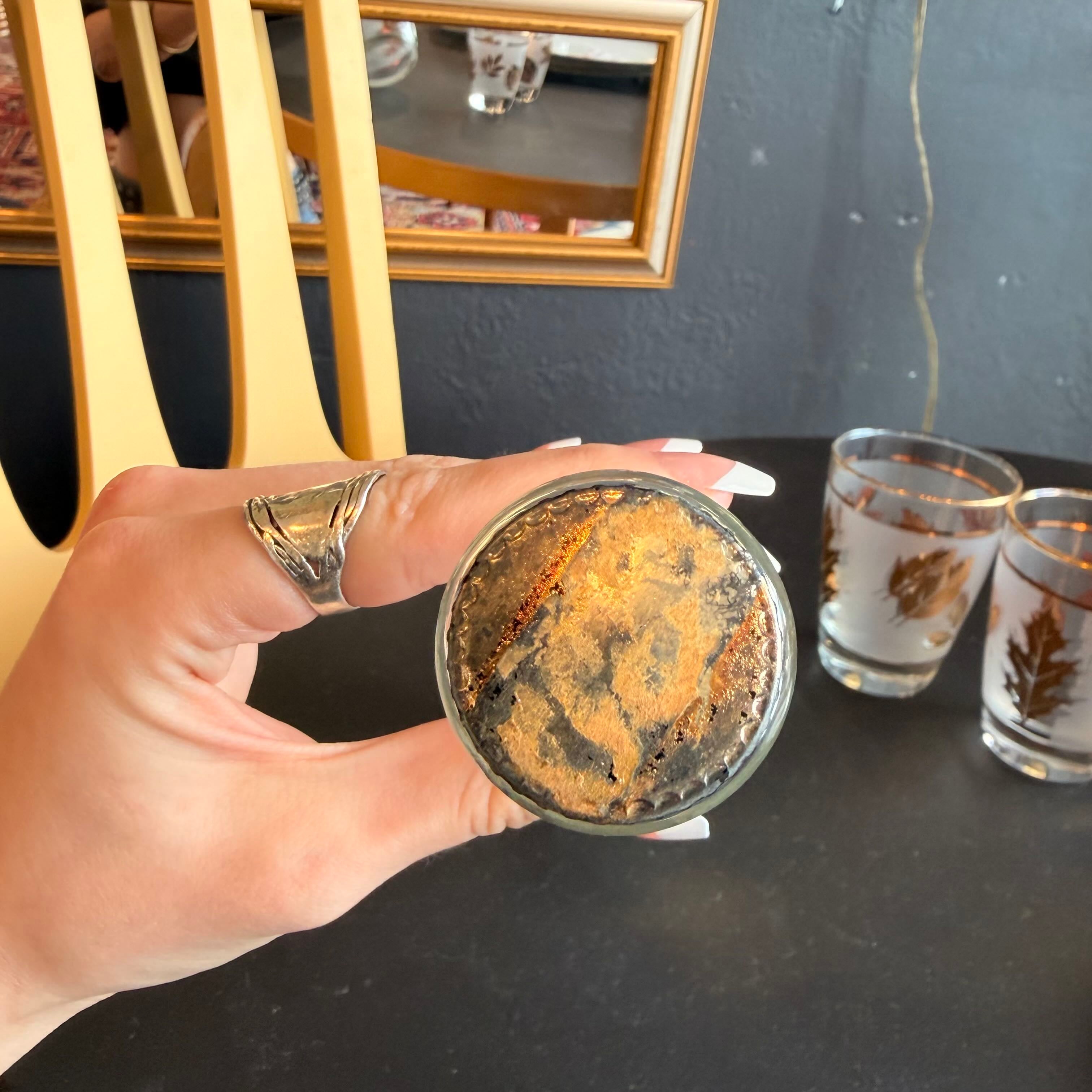 A hand holds a small clear glass tumbler filled with a dark brown textured substance, likely coffee grounds or tea, with fine golden-brown powder dusted on top. Two more similar glasses sit on a dark tabletop in the background, with a mirror and wall visible behind them.
