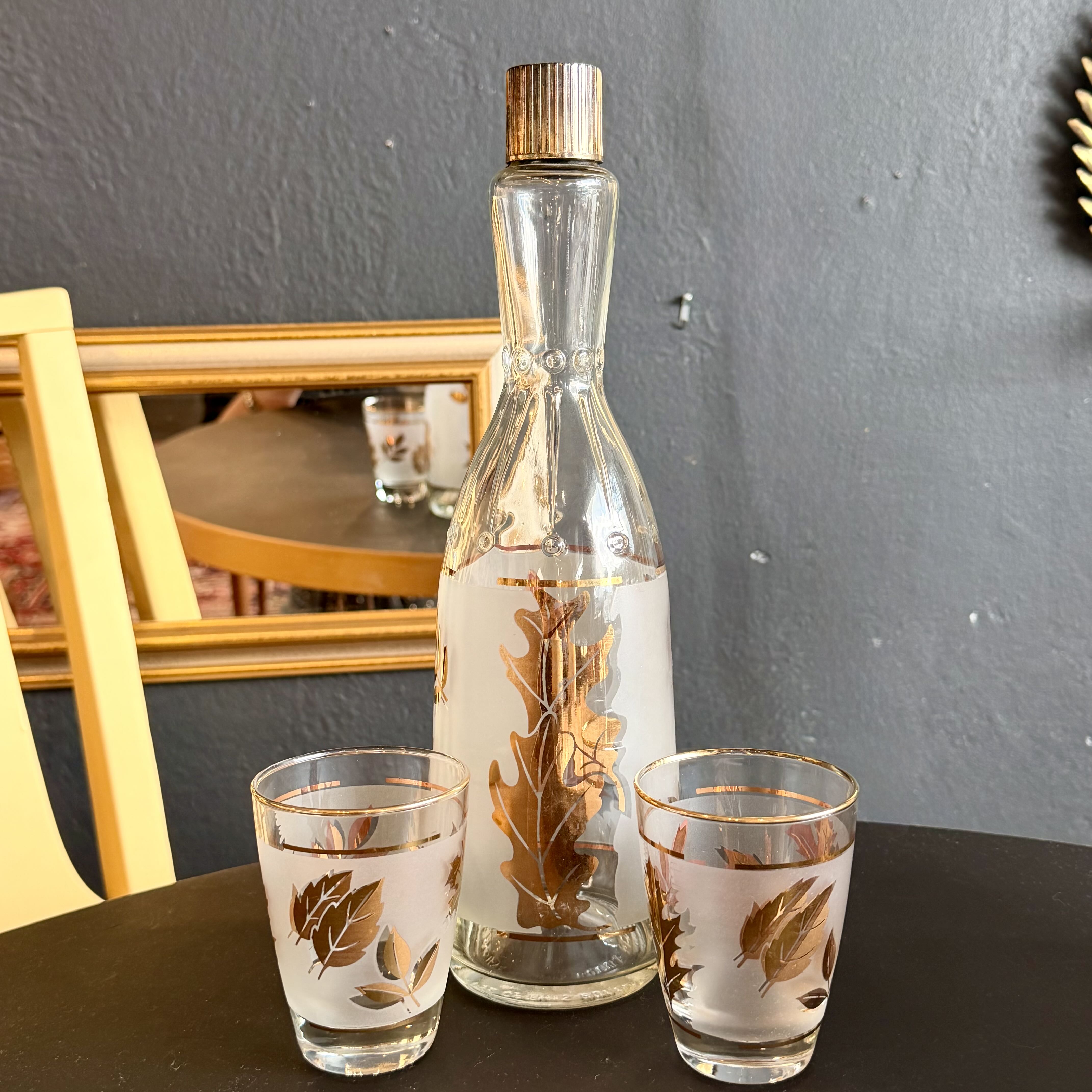 A clear glass bottle with a gold cap and gold leaf pattern, displayed on a dark table alongside two matching clear glass tumblers with gold leaf designs, set against a gray wall and yellow chair.