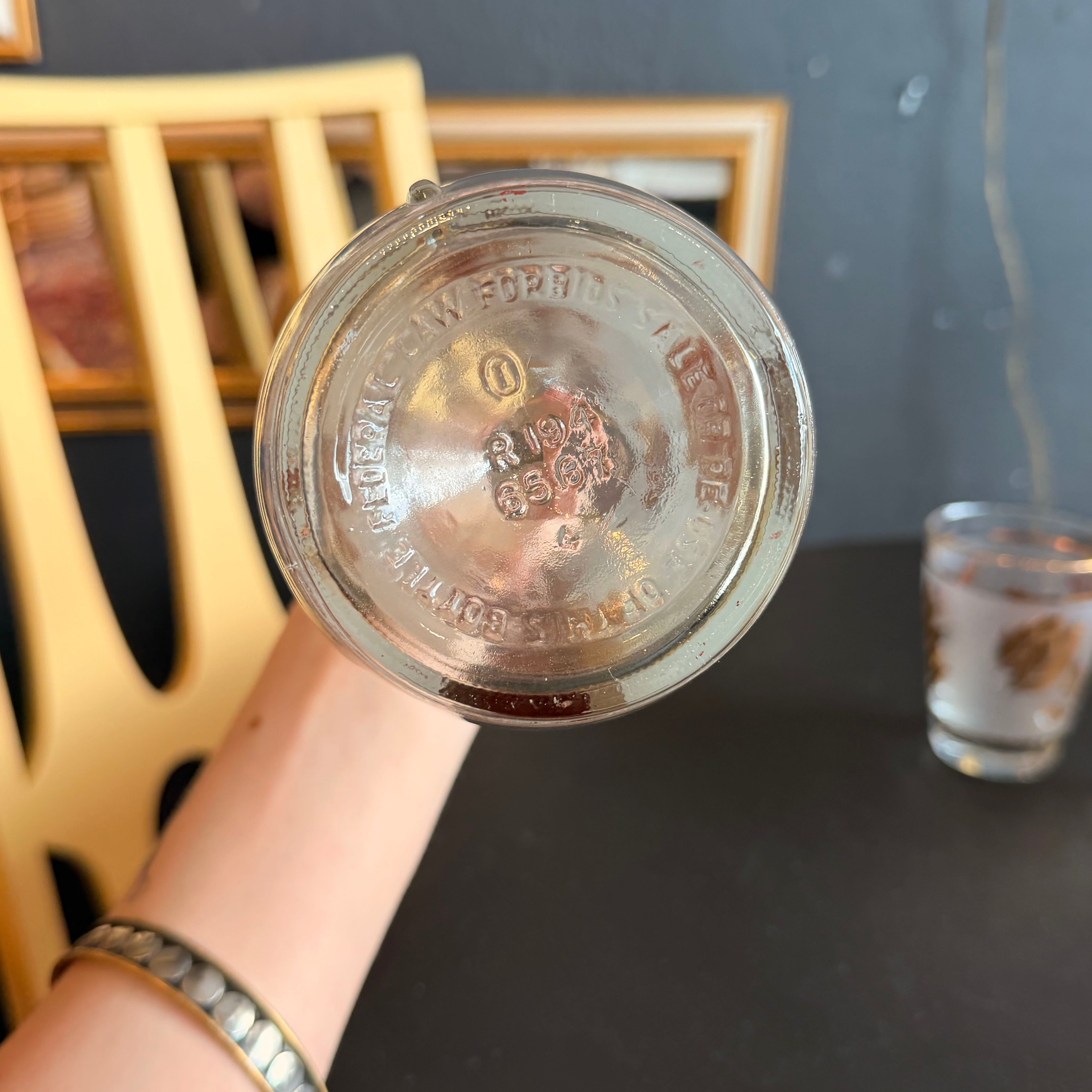 A hand holding the bottom of a clear glass jar or bottle, showing embossed text on the glass. A wooden chair and a dark table are in the background, along with a small shot glass.